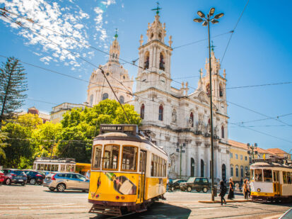 Eine wunderschöne belebte bunte Straße in Lissabon Portugal. Im Hintergrund zu sehen ist die prächtige Basilica da Estrela (Kirche) . Sie wird umringt von grünen Bäumen und den für Lissabon typischen Straßenbahnen.