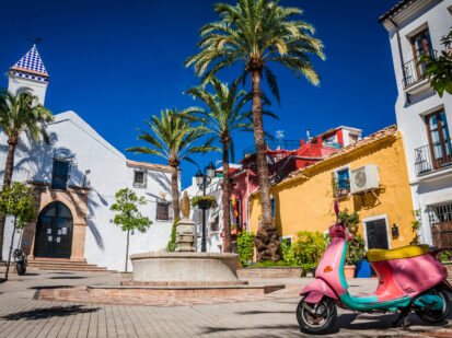 Ein schöner kleiner Platz in der Altstadt Marbella, mit weißen und bunten Häuschen. Inmitten des Platzes befindet sich ein kleiner Brunnen der von Palmen umzingelt ist. Vor dem Platz steht ein farbenfroher Roller, der das ganze Bild verschönert.