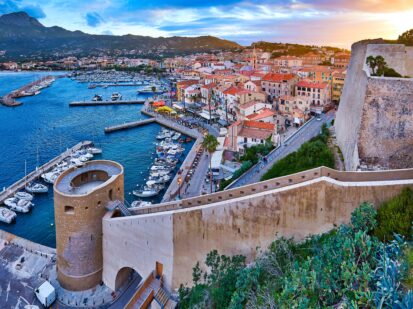 Blick von den Mauern der Zitadelle von Calvi auf die Altstadt mit historischen Gebäuden bei abendlichem Sonnenuntergang. Bucht mit Yachten und Booten. Luxuriöser Yachthafen.